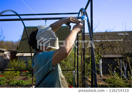 Against the background of a blue sky, a man welds metal pipes on his site. A man is assembling a metal base for a greenhouse, fastening pipes by welding. Metal welding concept. 116254310
