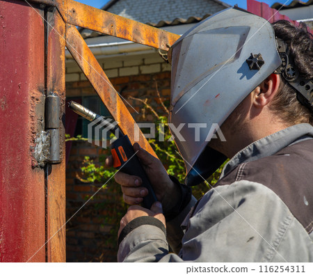 A man welds a metal gate using a welding machine. A man in a protective mask welds gate hinges. Repairing welds using a welding machine. A man welds a metal gate using a welding machine. A man in a protective mask welds gate hinges. Repairing welds using a welding machine. 116254311