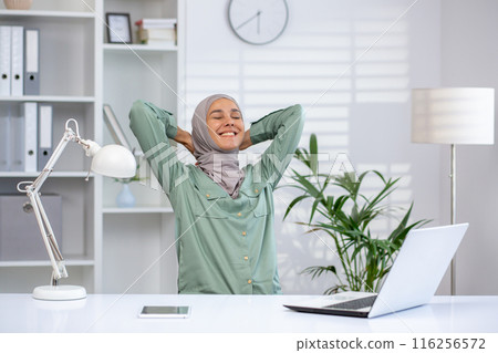A woman wearing a hijab relaxes and stretches at her office desk, smiling and enjoying a break. The modern workspace includes a laptop, tablet, desk lamp, and sleek decor, conveying comfort A woman wearing a hijab relaxes and stretches at her office desk, smiling and enjoying a break. The modern workspace includes a laptop, tablet, desk lamp, and sleek decor, conveying comfort 116256572