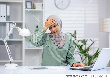 Stressed businesswoman wearing a hijab, holding her head while working on a laptop in a modern office environment. The imagery highlights concepts of workplace stress, concentration, and dedication. 116256581