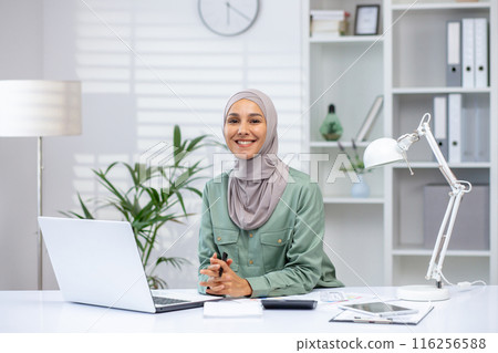 Confident businesswoman in a hijab sitting at her office desk with a laptop, smiling at the camera. The modern office environment is bright and organized, giving a professional and welcoming feel. Confident businesswoman in a hijab sitting at her office desk with a laptop, smiling at the camera. The modern office environment is bright and organized, giving a professional and welcoming feel. 116256588