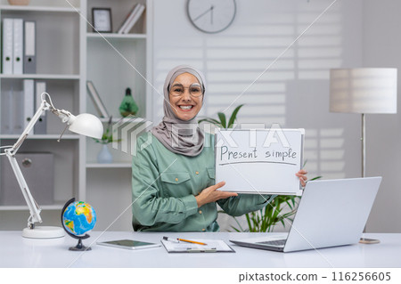 Female teacher wearing a hijab and glasses, conducting an online class on the present simple tense. Seated at the desk in a home office with a laptop, whiteboard, and educational materials. Female teacher wearing a hijab and glasses, conducting an online class on the present simple tense. Seated at the desk in a home office with a laptop, whiteboard, and educational materials. 116256605