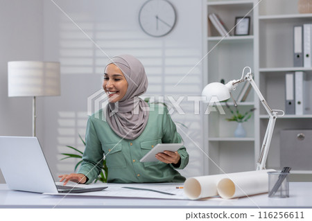 A smiling businesswoman in a hijab working at a modern office. She is using a laptop and holding a tablet, looking confident and professional. The environment is organized and conducive 116256611