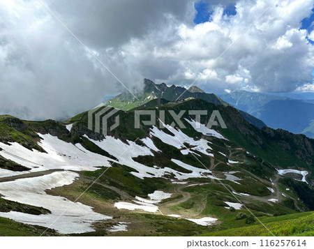 View of the Mountains landscape. Green mountain slopes with snow. Grassy field and rolling hills View of the Mountains landscape. Green mountain slopes with snow. Grassy field and rolling hills 116257614