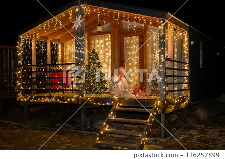Woman by Christmas tree. Festive setting. Illustrating holiday cheer and celebration. She is posing for a picture, and there are presents nearby, adding to the festive atmosphere. 116257899