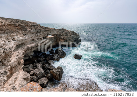 The ocean is calm and the sky is cloudy. The water is a deep blue color. The rocks are jagged and the waves are small. The ocean is calm and the sky is cloudy. The water is a deep blue color. The rocks are jagged and the waves are small. 116257928