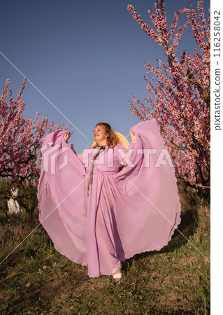 Woman blooming peach orchard. Against the backdrop of a picturesque peach orchard, a woman in a long pink dress and hat enjoys a peaceful walk in the park, surrounded by the beauty of nature. 116258042