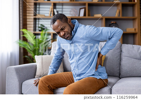 A man is sitting on a couch in a modern home interior, holding his back and expressing discomfort. The image captures the concept of back pain, discomfort, and health issues within a home setting. A man is sitting on a couch in a modern home interior, holding his back and expressing discomfort. The image captures the concept of back pain, discomfort, and health issues within a home setting. 116259964