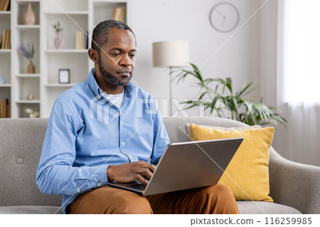 A man working from home on his laptop while sitting on a comfortable sofa in a well-decorated living room. The setting is relaxed, promoting productivity and focus. A man working from home on his laptop while sitting on a comfortable sofa in a well-decorated living room. The setting is relaxed, promoting productivity and focus. 116259985