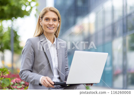Professional businesswoman in a gray suit using a laptop and headphones while sitting outdoors in an urban environment. She appears confident and focused, representing modern business work Professional businesswoman in a gray suit using a laptop and headphones while sitting outdoors in an urban environment. She appears confident and focused, representing modern business work 116260008