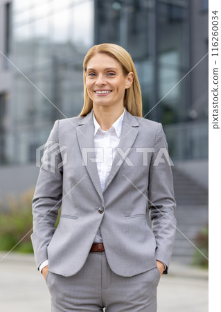 A confident businesswoman wearing a gray suit smiles while standing outdoors in front of a modern glass building. The professional look and positive expression evoke themes of success 116260034