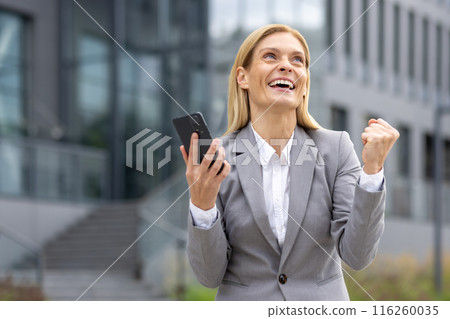 A joyful businesswoman in a gray suit celebrates success while holding a smartphone outdoors. She exudes confidence and happiness, illustrating achievement and professional triumph A joyful businesswoman in a gray suit celebrates success while holding a smartphone outdoors. She exudes confidence and happiness, illustrating achievement and professional triumph 116260035