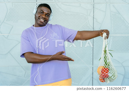 Portrait of happy young black man showing mesh bag of fresh groceries he bought at local market. Eco friendly (zero waste) lifestyle and healthy diet concept. 116260677