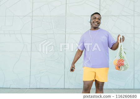 Sustainability, food shopping and eco friendly concept. Happy smiling African man listening music with headphones showing reusable net tote with fruits and vegetables on grey background. 116260680