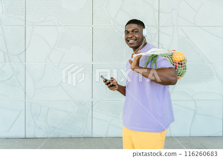 Black millennial hipster man wearing headphones using smartphone walking against wall carrying vegetables in reusable cotton mesh shopping bag, living green lifestyle. Black millennial hipster man wearing headphones using smartphone walking against wall carrying vegetables in reusable cotton mesh shopping bag, living green lifestyle. 116260683