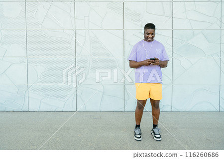Cheerful young man in sportwear using smartphone while stands against white wall urban background. Full height portrait Cheerful young man in sportwear using smartphone while stands against white wall urban background. Full height portrait 116260686