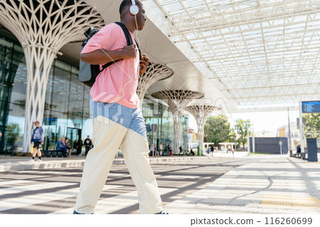 African man tourist or student in a pink shirt and blue jeans walks down a sidewalk, cross road with crosswalk with backpack. He is listening to music on his headphones 116260699
