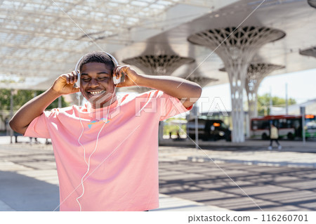 A man wearing a pink shirt is listening to music with headphones. He is smiling and he is enjoying himself. The scene takes place in an urban setting, with a bus. 116260701
