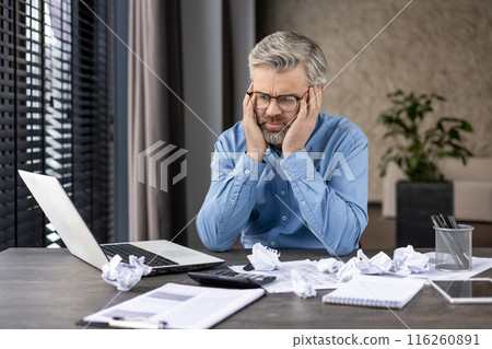 Middle-aged businessman feeling stressed and frustrated, sitting at his office desk with a laptop and crumpled papers. Concepts of stress, frustration, business problems, and workplace challenges. Middle-aged businessman feeling stressed and frustrated, sitting at his office desk with a laptop and crumpled papers. Concepts of stress, frustration, business problems, and workplace challenges. 116260891