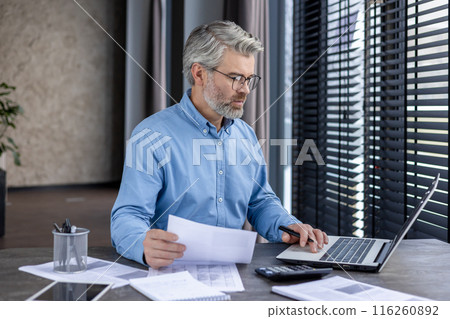 Mature businessman wearing glasses, working on laptop at a desk with documents and calculator in a modern office. Focused professional analyzing data, financial reports, and doing paperwork. Mature businessman wearing glasses, working on laptop at a desk with documents and calculator in a modern office. Focused professional analyzing data, financial reports, and doing paperwork. 116260892