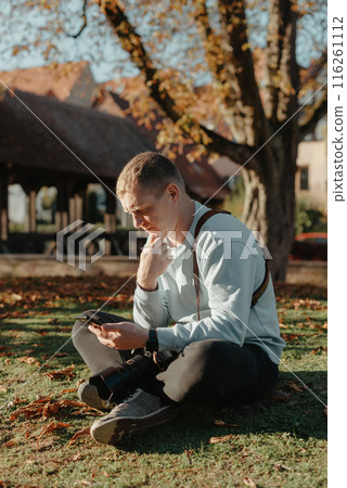 Professional Photographer Taking Picture Of Beautiful Autumn Park. Man Professional Photographer Sit With Camera And With Smartphone In Autumn Park. Retouched, Vibrant Colors, Brownish Tones. Professional Photographer Taking Picture Of Beautiful Autumn Park. Man Professional Photographer Sit With Camera And With Smartphone In Autumn Park. Retouched, Vibrant Colors, Brownish Tones. 116261112