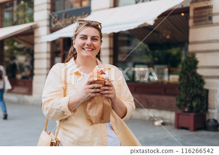 Beautiful overweight brunette 30s girl posing on the street, she has big croissant meal in hands. Unhealthy fattening food,high-calorie snack, eating on the go, take-out meals. Beautiful overweight brunette 30s girl posing on the street, she has big croissant meal in hands. Unhealthy fattening food,high-calorie snack, eating on the go, take-out meals. 116266542