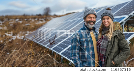 Couple in winter clothing standing next to solar panels in a field. Outdoor portrait for renewable energy and environmental awareness concepts. Winter landscape photography. Banner with copy space 116267516