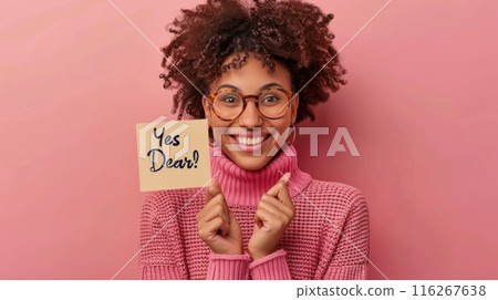 Happy Woman Holding Sign with Yes Dear. Cheerful woman in glasses and pink sweater, smiling broadly while holding a sign with "Yes Dear!" against a pink background. 116267638