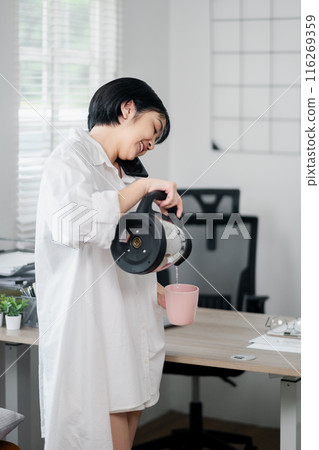 A woman in a white shirt pours coffee into a pink mug in a modern office with minimalist decor and natural light. A woman in a white shirt pours coffee into a pink mug in a modern office with minimalist decor and natural light. 116269359