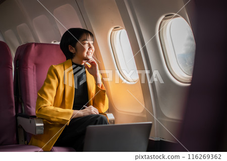 A woman in a yellow blazer sits by the airplane window, smiling and enjoying the view during her flight. A woman in a yellow blazer sits by the airplane window, smiling and enjoying the view during her flight. 116269362