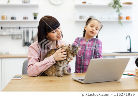 Mother and daughter with cat watching video on laptop in kitchen Mother and daughter with cat watching video on laptop in kitchen 116271959
