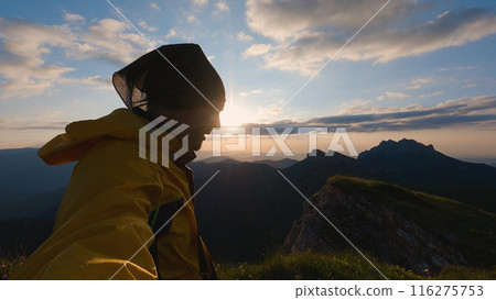 A man in a yellow windbreaker and head baffle stands against a mountainous landscape and watches the sunset. The serene beauty of nature is captured in this serene evening scene. 116275753