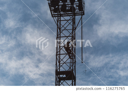 A silhouette of a person climbing a tall, metal tower against a dramatic, partly cloudy sky with light streaming through. The image evokes themes of adventure, risk-taking, and technology. 116275765
