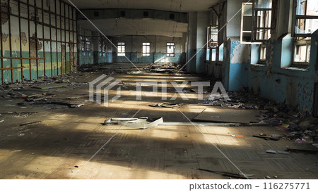 The interior of an abandoned dilapidated building with broken windows, peeling paint and rubbish scattered on the floor. Sunlight streams through the windows, casting shadows across the room. 116275771