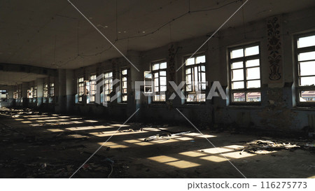 Abandoned dilapidated building with broken windows and debris strewn about. Sunlight streams in through the windows, casting long shadows across the floor. The walls show signs of decay and neglect. Abandoned dilapidated building with broken windows and debris strewn about. Sunlight streams in through the windows, casting long shadows across the floor. The walls show signs of decay and neglect. 116275773