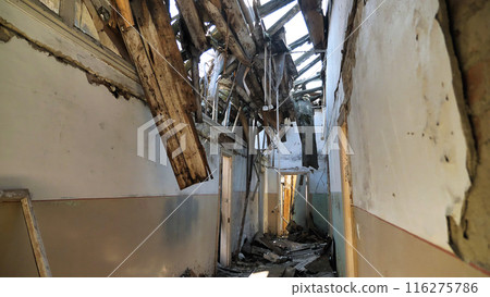 A dilapidated corridor with a badly damaged roof. The ceiling has collapsed, exposing wooden beams and debris scattered on the floor. Walls cracked and peeling, significant structural damage. A dilapidated corridor with a badly damaged roof. The ceiling has collapsed, exposing wooden beams and debris scattered on the floor. Walls cracked and peeling, significant structural damage. 116275786