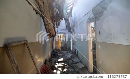 A dilapidated hallway with a collapsed ceiling and debris scattered on the floor. Sunlight streams through broken roof, illuminating the damaged walls and doorways. The scene abandonment and decay. 116275787