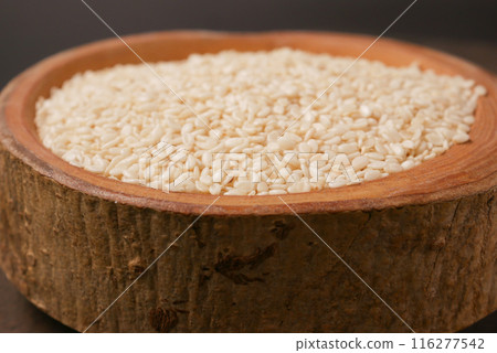 A wooden bowl filled with white sesame seeds, accompanied by a spoon, set on a dark surface 116277542