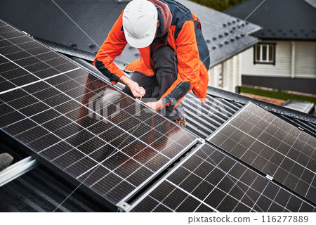 Man installer mounting photovoltaic solar panels on roof of house. Engineer in helmet installing solar module system with help of hex key. Concept of alternative, renewable energy. Man installer mounting photovoltaic solar panels on roof of house. Engineer in helmet installing solar module system with help of hex key. Concept of alternative, renewable energy. 116277889