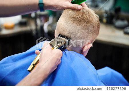 Back view of hairdresser shaving boy's head. Competent barber using electric shaver. Little client getting stylish men's haircut. 116277896