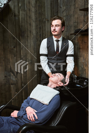 Experienced hairdresser with a french mustache washing client's hair. Professional hair cleaning in barbershop. 116277908