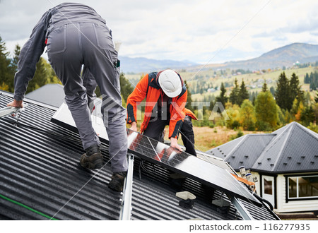 Installers building photovoltaic solar module station on roof of house. Men electricians in helmets installing solar panel system outdoors. Concept of alternative and renewable energy. 116277935