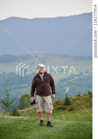 Male tourist hiking in the mountains, traveling in summer. Man standing on hill, holding camera, taking photo, admiring landscape, enjoying. Concept of tourism and adventure. 116277954