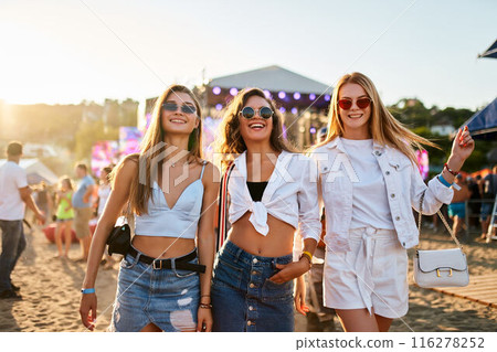 Group of joyful young women at sunset beach music festival. Friends dance, enjoy live band, summer vibes by sea. Smiling girls with sunglasses, casual fashion at outdoor concert, vacation leisure. Group of joyful young women at sunset beach music festival. Friends dance, enjoy live band, summer vibes by sea. Smiling girls with sunglasses, casual fashion at outdoor concert, vacation leisure. 116278252