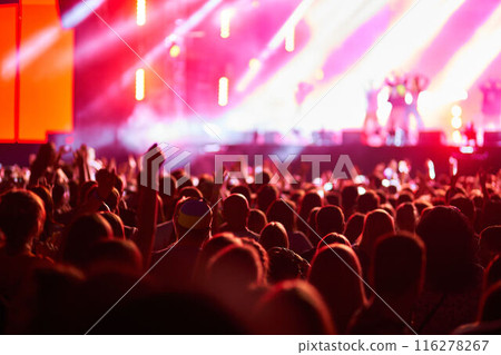 Concertgoers enjoy live music at outdoor fest, vibrant stage lights illuminate performers. Fans wave hands, dance, celebrate. Summer event vibe, festival season, night entertainment atmosphere. 116278267