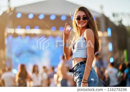 Joyful young woman enjoys summer music festival on beach, waves hand sign with stage lights, crowd in background. Trendy outfit, sunglasses, vibrant atmosphere at coastal event for promo. Joyful young woman enjoys summer music festival on beach, waves hand sign with stage lights, crowd in background. Trendy outfit, sunglasses, vibrant atmosphere at coastal event for promo. 116278296