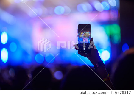 Concertgoer films live music event with smartphone, hand raised above crowd, capturing stage lights, performers in blurred background. Energetic festival atmosphere, fans enjoy night performance. 116278317