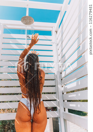A woman in a bikini stands in front of a white shower stall. She is smiling and she is enjoying the moment. 116279481
