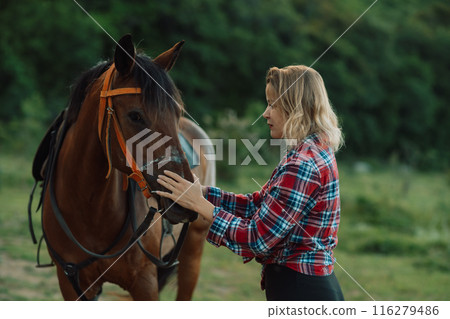 Happy blonde with horse in forest. Woman and a horse walking through the field during the day. Dressed in a plaid shirt and black leggings. Happy blonde with horse in forest. Woman and a horse walking through the field during the day. Dressed in a plaid shirt and black leggings. 116279486