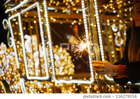 Woman holding sparkler night while celebrating Christmas outside. Dressed in a fur coat and a red headband. Blurred christmas decorations in the background. Selective focus 116279558